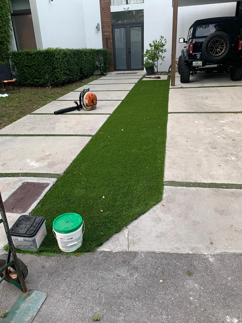 Artificial turf installation in progress between concrete driveway slabs, with tools and materials still on site