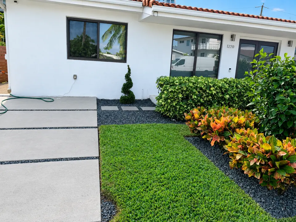 Evenly lit front garden with green lawn, croton planting, dark stone beds, and concrete driveway slabs beside a white home