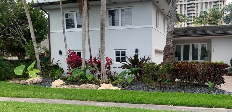 Finished tropical garden with palms, colorful foliage, natural stone, and a neatly edged lawn around a white home